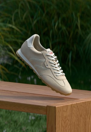 Zapatilla deportiva beige con material de ante, cordones blancos, diseño suave, punta redondeada y branding sutil en el talón, mostrada sobre una superficie de madera.