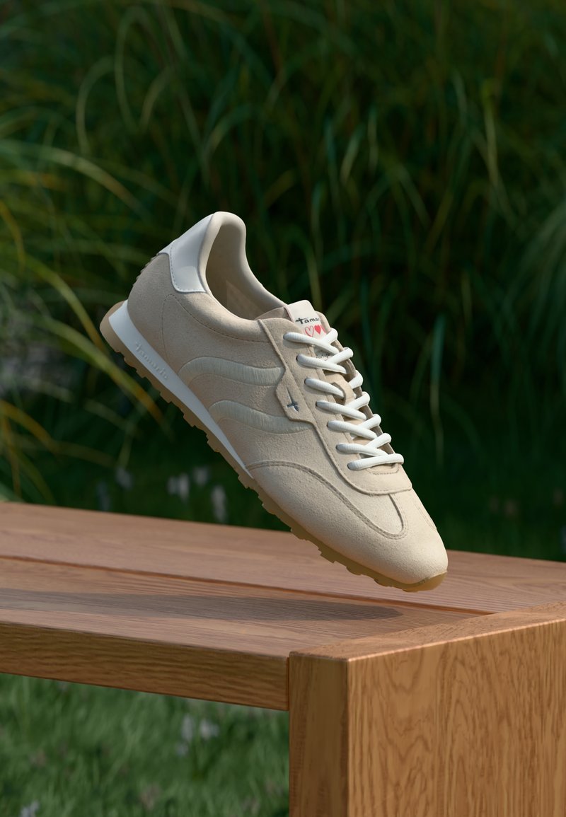 Chaussure de sport beige en daim, lacets blancs, design épuré, bout arrondi et branding discret au talon, présentée au-dessus d'une surface en bois.