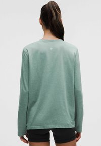 Top cu mânecă lungă de culoare verde pal, cu guler rotund, croială lejeră și textură subtilă. Prezintă un logo mic în partea superioară a spatelui.