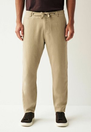 Beige bomuldsblandingsbukser med elastisk talje, sidelommer og lige pasform, parret med mørke sneakers på en ensfarvet baggrund.
