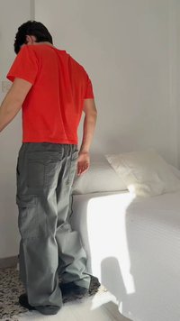 Persoon in een rood shirt en grijze cargobroek staat naast een wit bed met een kussen in een met zonlicht gevulde kamer.
