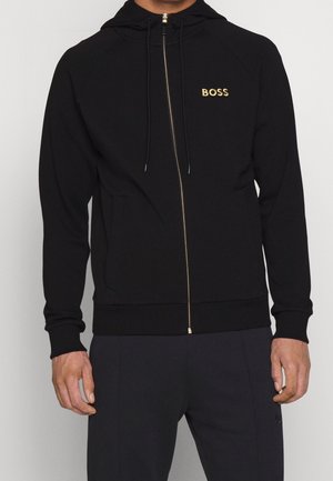 Homme portant un sweat à capuche noir à fermeture éclair avec le logo doré "BOSS" sur la poitrine et un pantalon assorti noir, debout devant un fond uni.