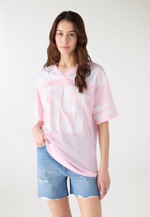 Giovane donna con lunghi capelli castani che indossa una maglia sportiva rosa chiaro in rete con il numero "89" in bianco e pantaloncini di jeans strappati, in piedi contro uno sfondo bianco.
