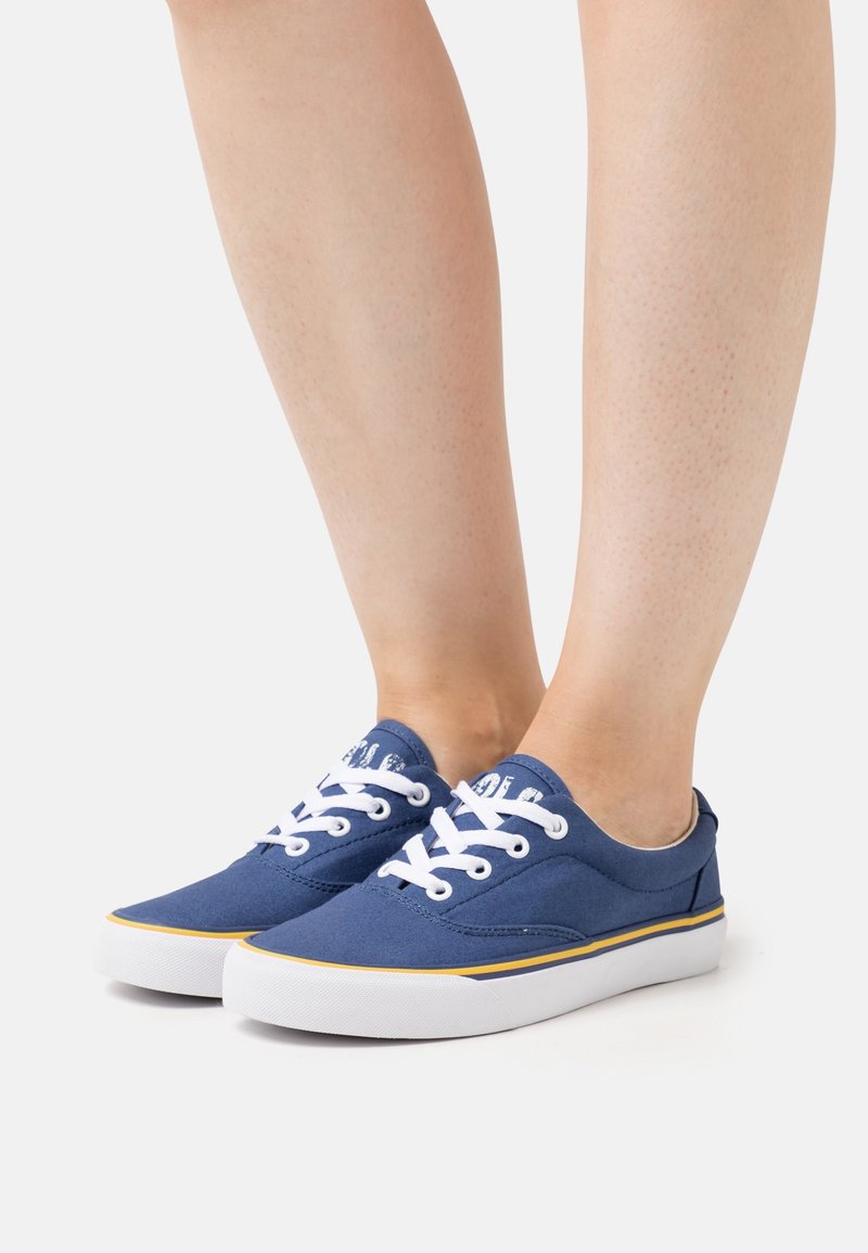 Jambes portant des baskets en toile bleue avec des lacets blancs, des semelles blanches et une bordure jaune sur un fond clair uni.