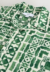 Chemise à manches courtes avec un motif géométrique vert et blanc, fabriquée en tissu léger, avec un col à rabat et une fermeture à boutons.
