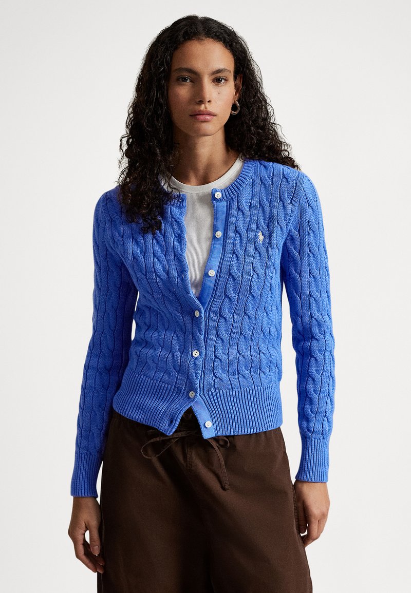 Zila kabeladīta kardigāna ar apaļu kakla izgriezumu, pogu aizdari un ribotu apakšmalu. Valkāta pār gaišu topu un kombinēta ar brūnām biksēm.