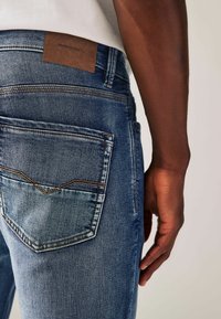Sinised denim teksad nõrga viimistlusega, millel on tagatasku dekoratiivse tikandiga ja pruun nahklabel vöökoha juures.