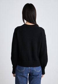 Maglione nero a maglia con scollatura e bordo a costine, dotato di maniche lunghe e una vestibilità morbida. Indossato con jeans blu.