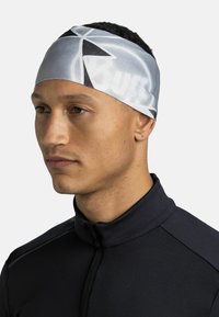 Diadema en gris con un patrón geométrico, fabricada con un tejido suave y elástico; llevada por una persona con una camiseta de cremallera negra.