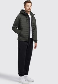 Dunkelgrüne gepolsterte Jacke mit Reißverschluss vorne, Kapuze und gestepptem Muster. Kombiniert mit schwarzen Jogginghosen und weißen Turnschuhen.