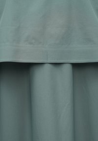 Vrstvené šaty z jemnej, svetlomodrozelenkastej látky. Horná časť je hladká, zatiaľ čo spodná má splývavú, nariasenú textúru s viditeľnými švami.