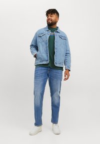 Lichtblauwe spijkerjack met twee borstzakken, gecombineerd met een donkergroen sweatshirt. Lichtblauwe jeans en witte sneakers maken de outfit compleet.