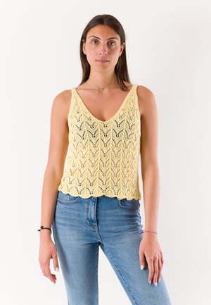 Donna con lunghi capelli scuri indossa un top giallo a maglia senza maniche e jeans blu, in piedi contro uno sfondo bianco semplice.