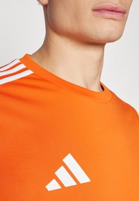 Orange athletic shirt made of smooth fabric, featuring a crew neck, three white stripes on one shoulder, and a large white triangular logo.