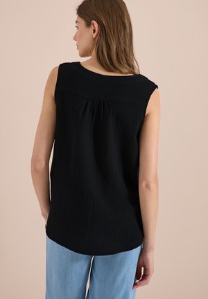 Femme aux longs cheveux bruns portant un top noir texturé sans manches et un jean bleu clair, debout devant un fond beige uni.
