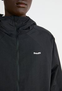 Black hooded jacket made of lightweight material with a smooth texture. Features a front zipper and a white logo on the chest.