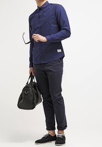 Marineblaue Langarmbluse mit weißen Punkten, kombiniert mit dunklen Hosen, einer schwarzen Tasche und schwarzen Schuhen mit weißen Schnürsenkeln.