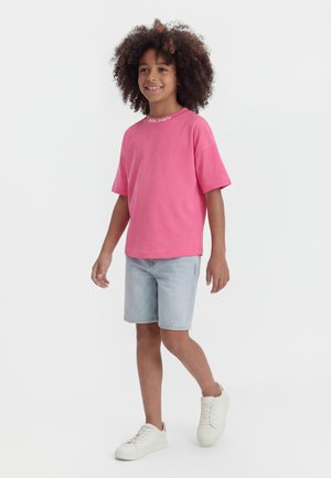 Dieťa s kučeravými vlasmi v ružovom tričku s krátkym rukávom, svetlých rifľových kraťasoch a bielych teniskách, kráčajúce a usmievajúce sa na jednofarebnom pozadí.