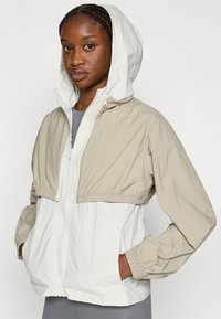 Ελαφρύ μπουφάν με φερμουάρ, με μπεζ επάνω μέρος και λευκό κάτω τμήμα, με ελαστικά μανσέτα, κουκούλα και πλαϊνές τσέπες. Υφή υφάσματος.