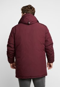 Giacca imbottita color marrone scuro con collo alto, cappuccio regolabile e cuciture visibili. Tessuto dalla texture liscia, design di media lunghezza e tasche laterali.