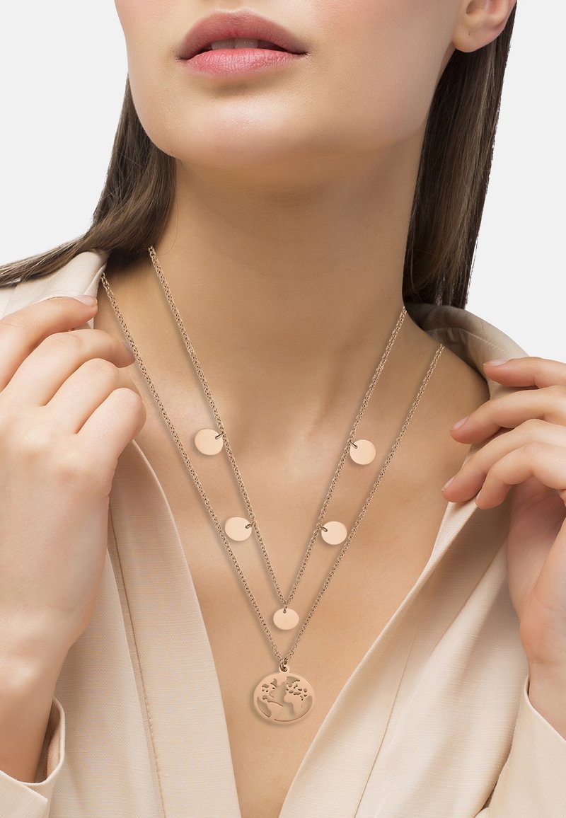 Gros plan sur une femme portant des colliers en or superposés avec des pendentifs ronds, dont un comportant une carte du monde découpée, vêtue d'un chemisier beige clair.