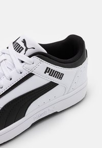 Baskets Puma présentant un design en cuir noir et blanc, des panneaux texturés, un col rembourré et une semelle en caoutchouc blanche. Logo affiché sur la languette.