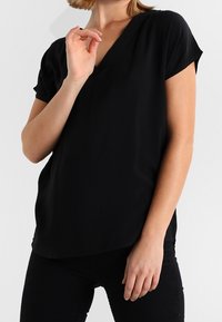 Top negro de cuello en V con mangas cortas, textura suave, corte regular y un dobladillo ligeramente curvado, combinado con pantalones negros.