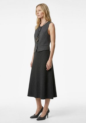 Nainen, joka pukeutuu hihattomaan harmaaseen napitettuun liiviin, pitkä tummaharmaa hame, mustat korkokengät ja pyöreä riipuskaulakoru, seisoo sivuttain.