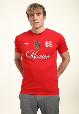 Jeune homme portant un maillot de football rouge du Mexique avec le numéro 86, le logo et la marque "TOFFS", debout les mains dans le dos.
