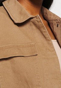 Tan denimjakke med krave, en brystlomme og en tekstureret overflade. Stoffet er robust og let børstet.