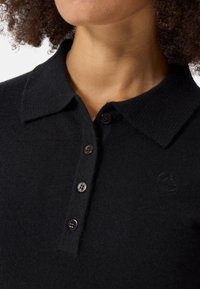 Svart stickad polo-shirt med krage, med en placket med tre knappar och en subtil logodetalj på bröstet.