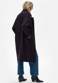 Lange, oversized jas van donkerpaars fabric met brede mouwen en een minimalistisch ontwerp. Gecombineerd met losse denim en zwarte haklaarzen.