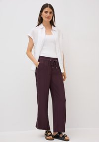 Witte linnen shirt met korte mouwen over een wit tanktop, gecombineerd met donkerpaarse, losse broek met een elastische tailleband en een trekkoord.