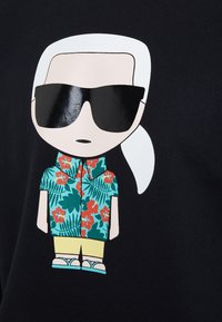 Camiseta gráfica con un personaje de dibujos animados que tiene el cabello blanco, gafas de sol, una camisa floral en turquesa y rojo, y pantalones cortos amarillos.