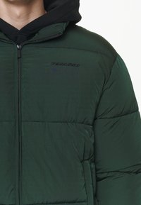 Chaqueta acolchada verde con cierre de cremallera, con sudadera con capucha negra debajo. Logo "PEGADOR" en negro en el pecho. Material texturizado.