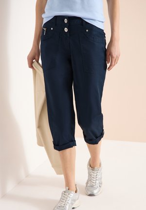 Personne portant un pantalon capri marine retroussé, des baskets argentées, un haut bleu clair, tenant une veste beige dans la main gauche sur un fond neutre.