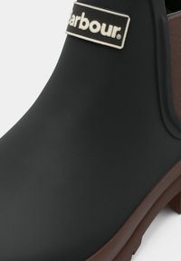Zwarte rubberen laars met een matte afwerking, voorzien van een bruine accent en een merklabel. Stevige zool en comfortabele vorm voor buitengebruik.