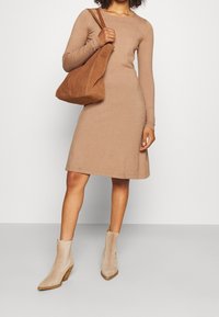 Abito a maniche lunghe, lungo fino al ginocchio, in morbido tessuto di maglia color marrone chiaro; abbinato a una borsa a tracolla in suede marrone e stivaletti beige con un design triangolare.