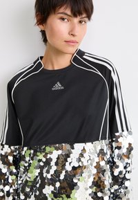 Jauns cilvēks ar īsiem brūniem matiem, tērpies melnā Adidas t-kreklā ar baltām svītrām un lieliem sudraba mirdzumiem apakšējā daļā.