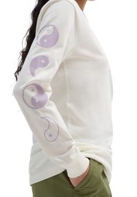 Person in einem weißen Langarmshirt mit lila Yin-Yang-Symbolen auf den Ärmeln und grüner Hose, Hand in der Tasche, Seitenansicht.