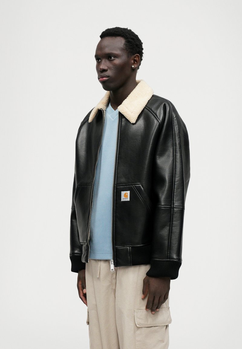 Schwarze Lederbomberjacke mit elfenbeinfarbenem Schafslederkragen, Rippbündchen und Reißverschluss. Getragen über einem hellblauen V-Ausschnitt-Shirt und beigen Hosen.