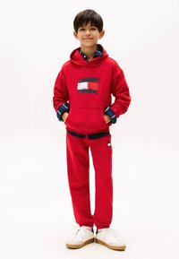 Rotes Kapuzen-Sweatshirt mit Logodesign auf der Brust, kombiniert mit passenden roten Jogginghosen. Die Sneaker haben ein weißes Obermaterial mit gumifarbenen Sohlen.