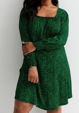Woman wearing a green and black patterned long-sleeve dress with square neckline, silver bracelet, and rings, standing against a plain background.