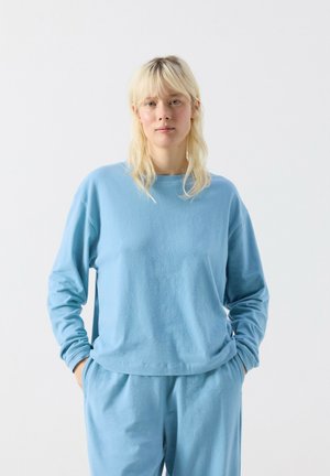 Frau mit blondem Haar, die einen hellblauen Langarmpullover und passende Hose trägt, steht mit den Händen in den Taschen vor einem weißen Hintergrund.