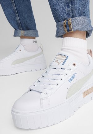 Białe sneakersy na platformie Puma z jasnobeżowymi detalami, noszone z białymi skarpetkami i podwiniętymi niebieskimi dżinsami na jasnym tle.