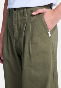 Persona che indossa pantaloni verde oliva con una mano nella tasca anteriore, mostrando delle pieghe e una piccola etichetta bianca sul bordo della tasca.