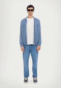 Jasnoniebieska bluza na zamek z logo, pod spodem biała koszulka, jasnoniebieskie jeansy i ciemne trampki. Model nosi czarne okulary przeciwsłoneczne.