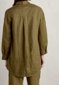 Olive green linen shirt with a back pleat, long sleeves, and buttoned cuffs. The hem is curved, and the fabric has a textured finish.