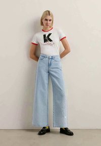 Biele tričko s krátkym rukávom a červenými akcentmi, s grafickým dizajnom. Svetlomodré široké nohavice s vyšitým textom na boku. Čierne topánky.