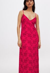 Robe maxi fuchsia avec de fines bretelles spaghetti et un décolleté en V, présentant des motifs floraux noirs sur un tissu transparent.